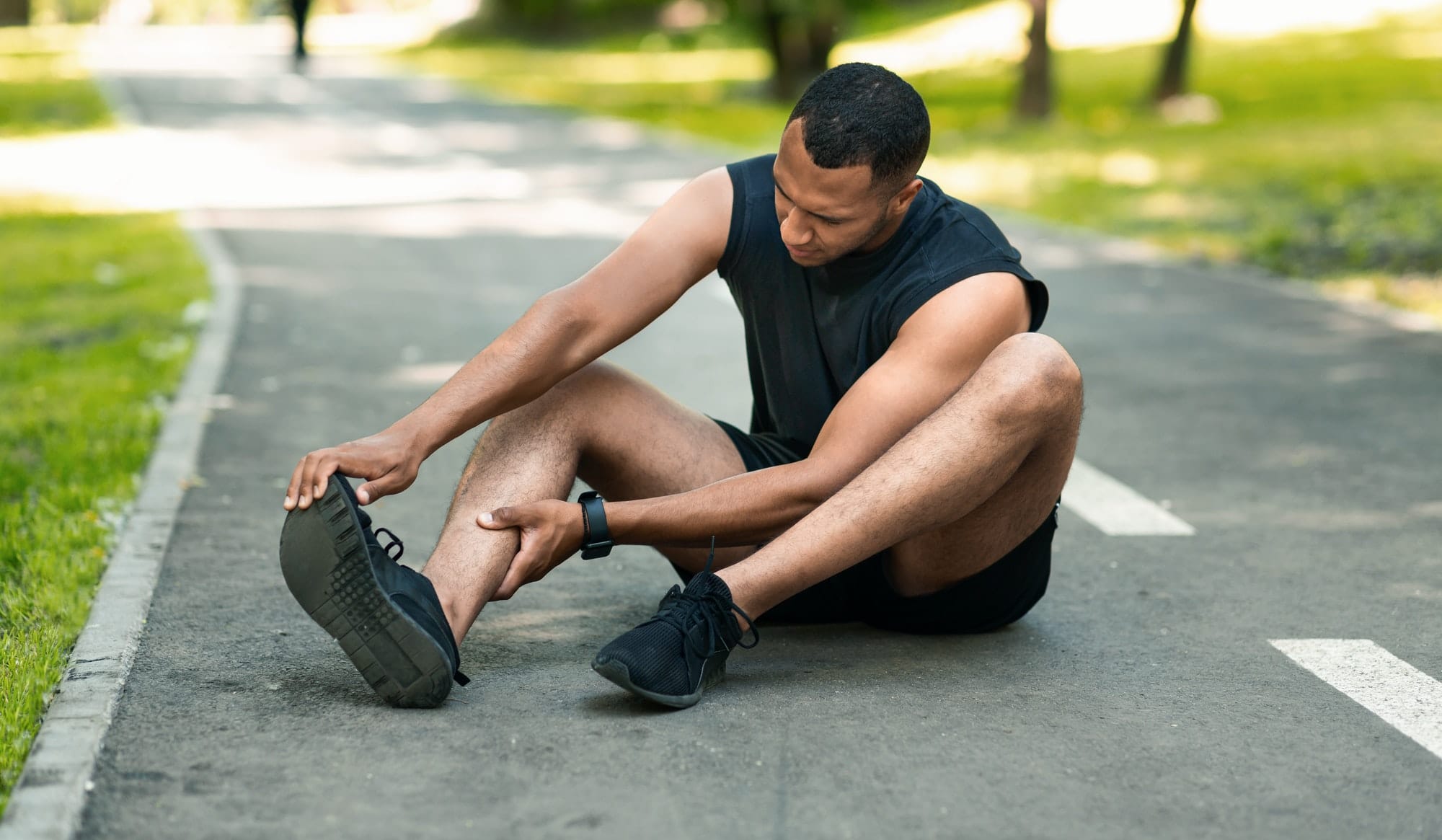 Sports injury. African American runner sitting on jogging track and feeling pain in his ankle