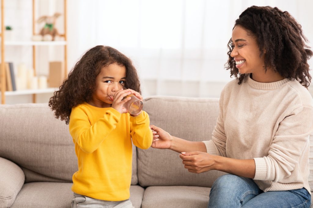 Mother Feeding Daughter, Caring For Kid Sitting On Couch Indoor