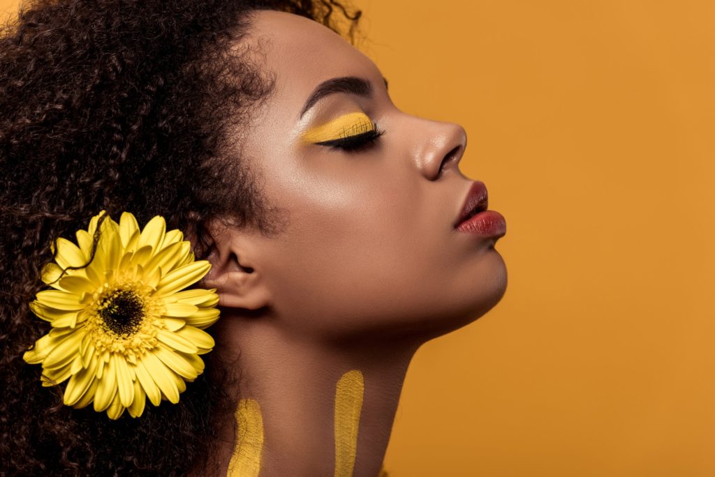 Stylish african american woman with artistic make-up and gerbera in hair dreaming isolated on orange