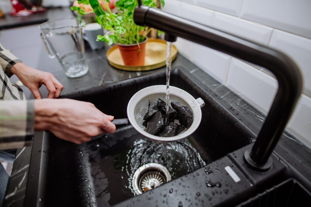 Woman cleaning shungite stones in sieve with pouring water in sink